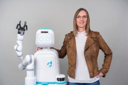 A woman puts her arm around the shoulder of a friendly-looking robot with one arm. The robot is a little shorter than a human.